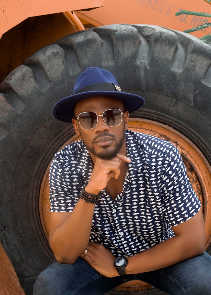 Home Fashionable portrait of a stylish man in blue hat and sunglasses posing outdoors with tractor tire backdrop.